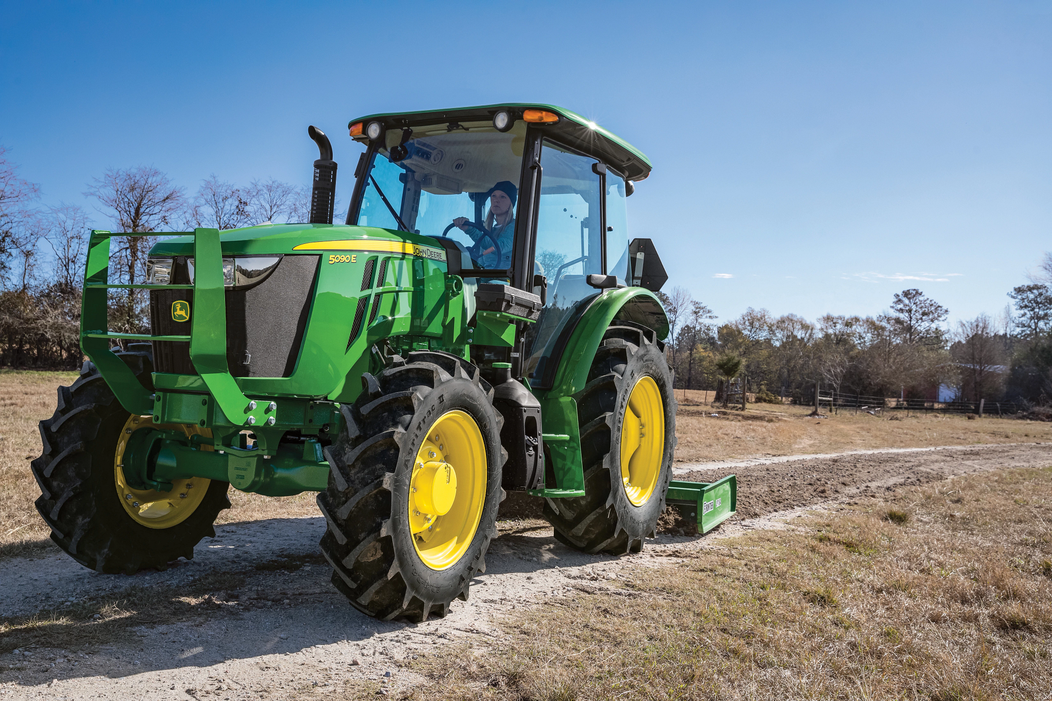 Green tractor with yellow wheels driven by a person travels along a gravel road in a rural landscape, with dry grass, scattered trees, and a clear blue sky in the background